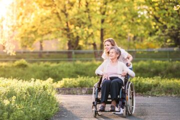 A woman accompanying an older adult in a wheelchair along a sunlit park path showcasing compassionate care